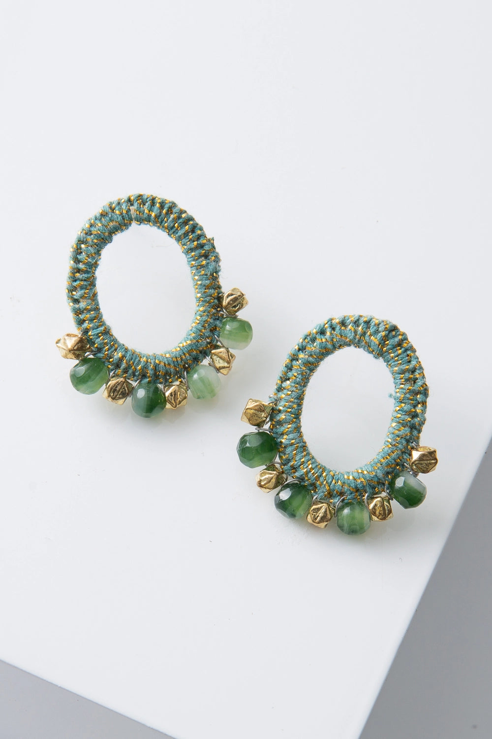 The Anjana Crochet Earrings are mid-size post earrings. The ear post connects to an oval hoop that is wrapped in mint green and metallic gold crocheted thread. Faceted gold metal beads and jade green glass beads are arranged along the bottom of the hoop.