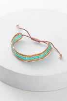 The Aqua Weave Bracelet is composed of blush colored thread and small glass beads. A row of turquoise colored beads is outlined with a row of gold beads on top and bottom. The bracelet can be adjusted by pulling on either end of the thread clasp.