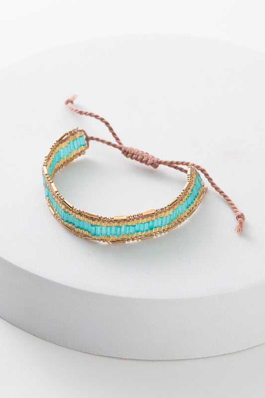 The Aqua Weave Bracelet is composed of blush colored thread and small glass beads. A row of turquoise colored beads is outlined with a row of gold beads on top and bottom. The bracelet can be adjusted by pulling on either end of the thread clasp.