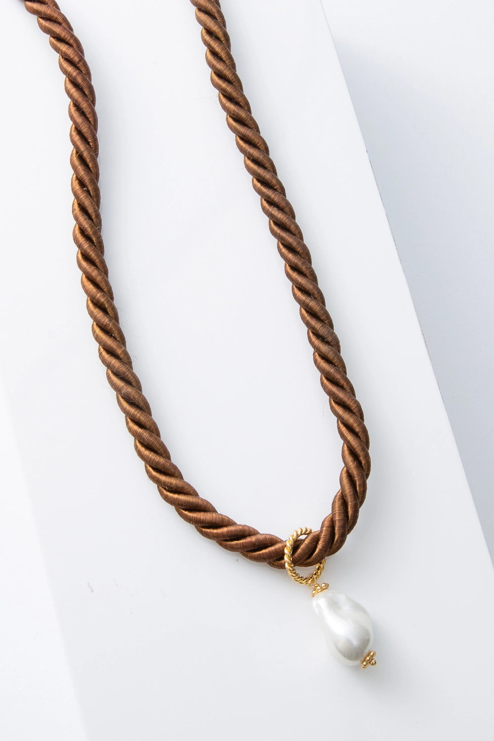 This is a rope-style necklace composed of twisted brown silk thread. At the base of the necklace, a single pearl is attached with a gold ring. The pearl has an irregular, organic shape.