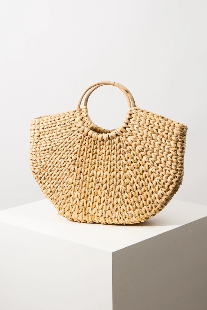 The Beach Basket Tote is a woven crescent-shaped bag composed entirely from natural grass. The grass has a knitted look and has a natural tan color. The grass is attached to two round cane handles.