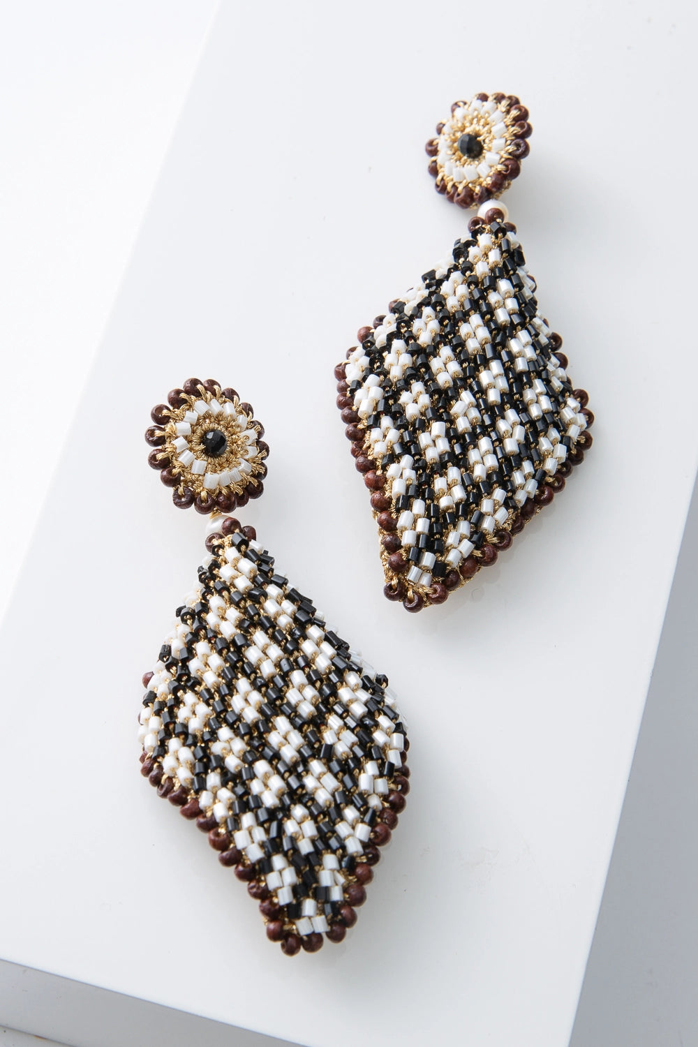 These are large, diamond-shaped beaded earrings. The posts are covered in gold wire and beads arranged to form a floral pattern, with a black bead at the center, a circle of white beads surrounding it, and a circle of brown beads around the outside. Hanging from the posts is a large diamond composed of the same materials. The beads are arranged to form a gingham-like black and white pattern, with brown beads surrounding the outside of the diamond.