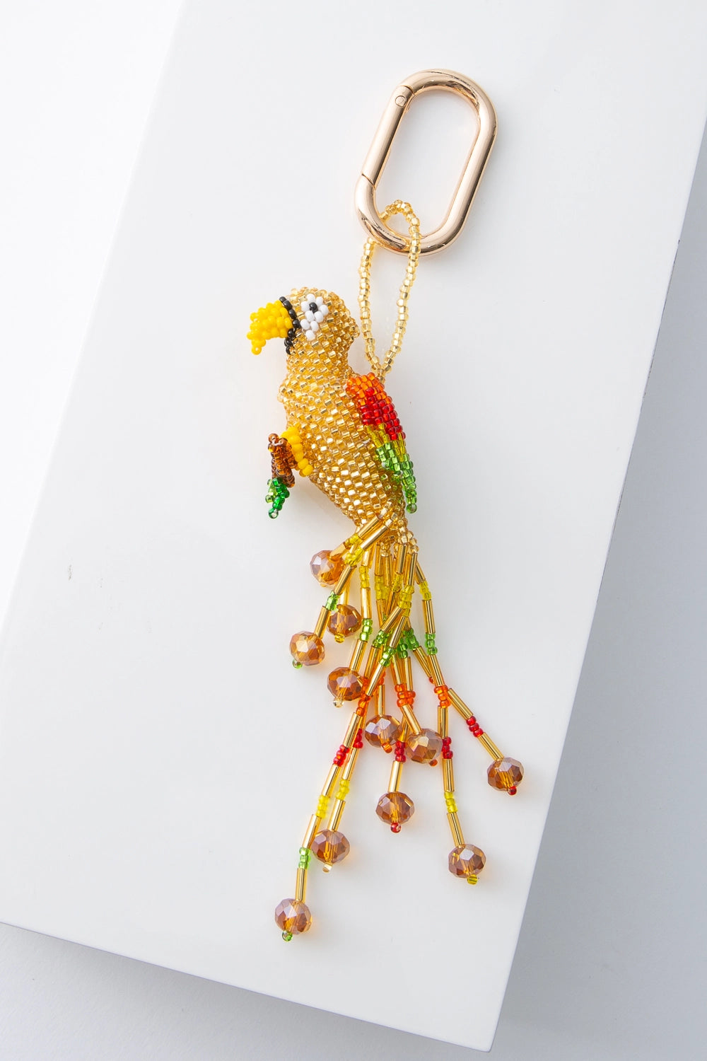 A three-dimensional beaded bird hangs from a brass oval keyring. The bird is primarily composed of translucent yellow beads. It has a beak, eyes, wings, and tail feathers composed of different colored beads. The beak is gold and the wings are red, yellow, and green. A long beaded fringe hangs down, forming the tail feathers.