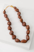 The Bisola Bauble Necklace features a single strand of shiny brown seeds called Bisola Seeds. The seeds each have their own unique shapes and natural striations. The seeds closest to the necklace clasps are the smallest, and they gradually increase in size towards the bottom of the necklace. The necklace is finished with a gold chain clasp.