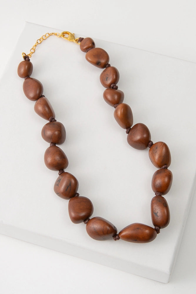 The Bisola Bauble Necklace features a single strand of shiny brown seeds called Bisola Seeds. The seeds each have their own unique shapes and natural striations. The seeds closest to the necklace clasps are the smallest, and they gradually increase in size towards the bottom of the necklace. The necklace is finished with a gold chain clasp.