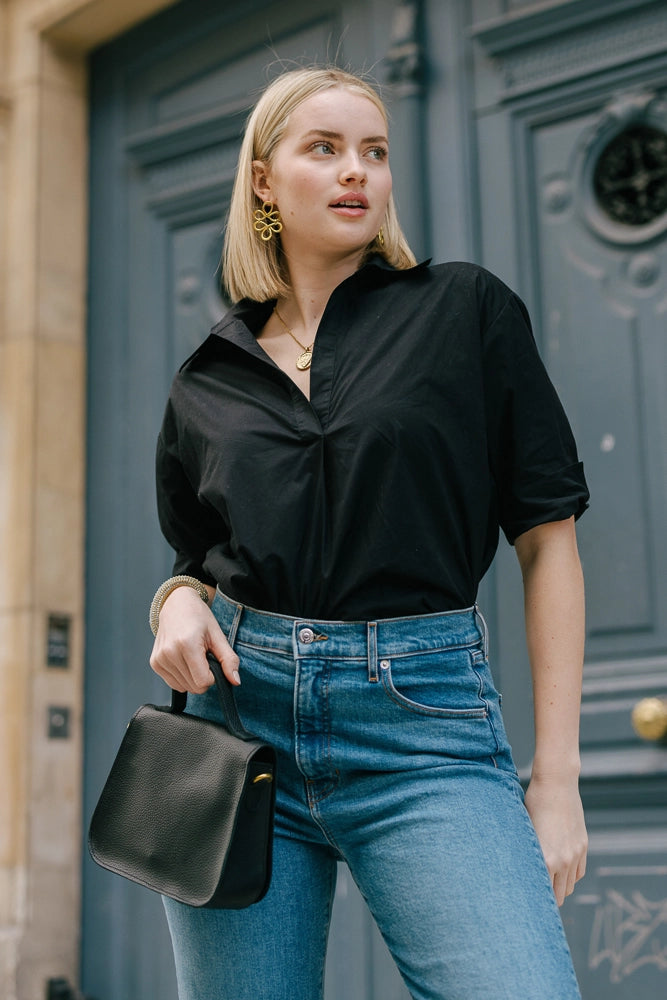 A model wears the Black Breezy Tunic tucked into jeans. It is a boxy black cotton tunic with cuffed sleeves that hit just above the elbow. It has an open v-neck and fold-down collar.