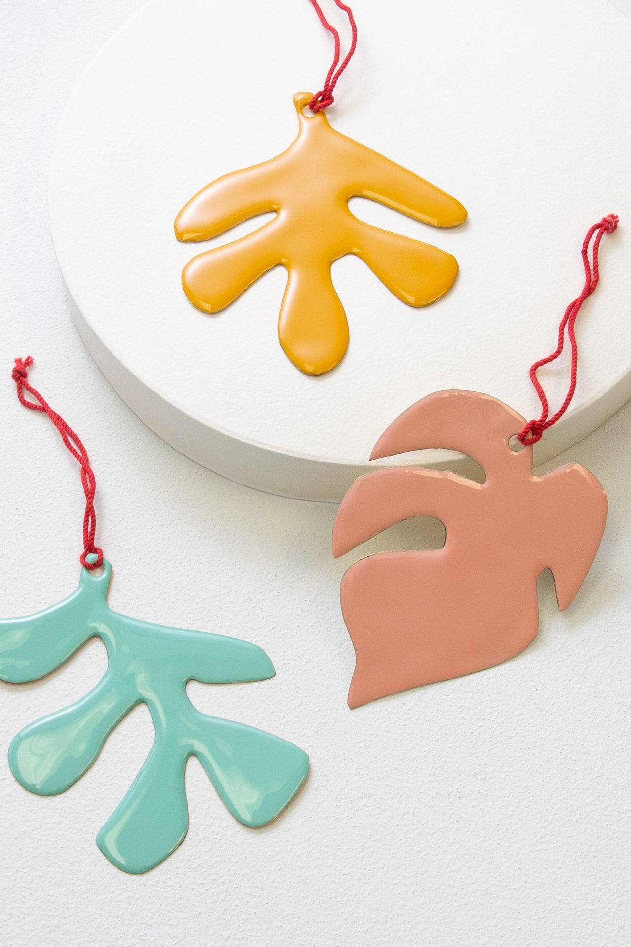 The three ornaments included in the Botanical Ornament set are composed of brass coated in glossy resin. Each is a different leaf shape and a different color. One is teal, one is gold, and one is peach. Each is finished with a red cord loop.