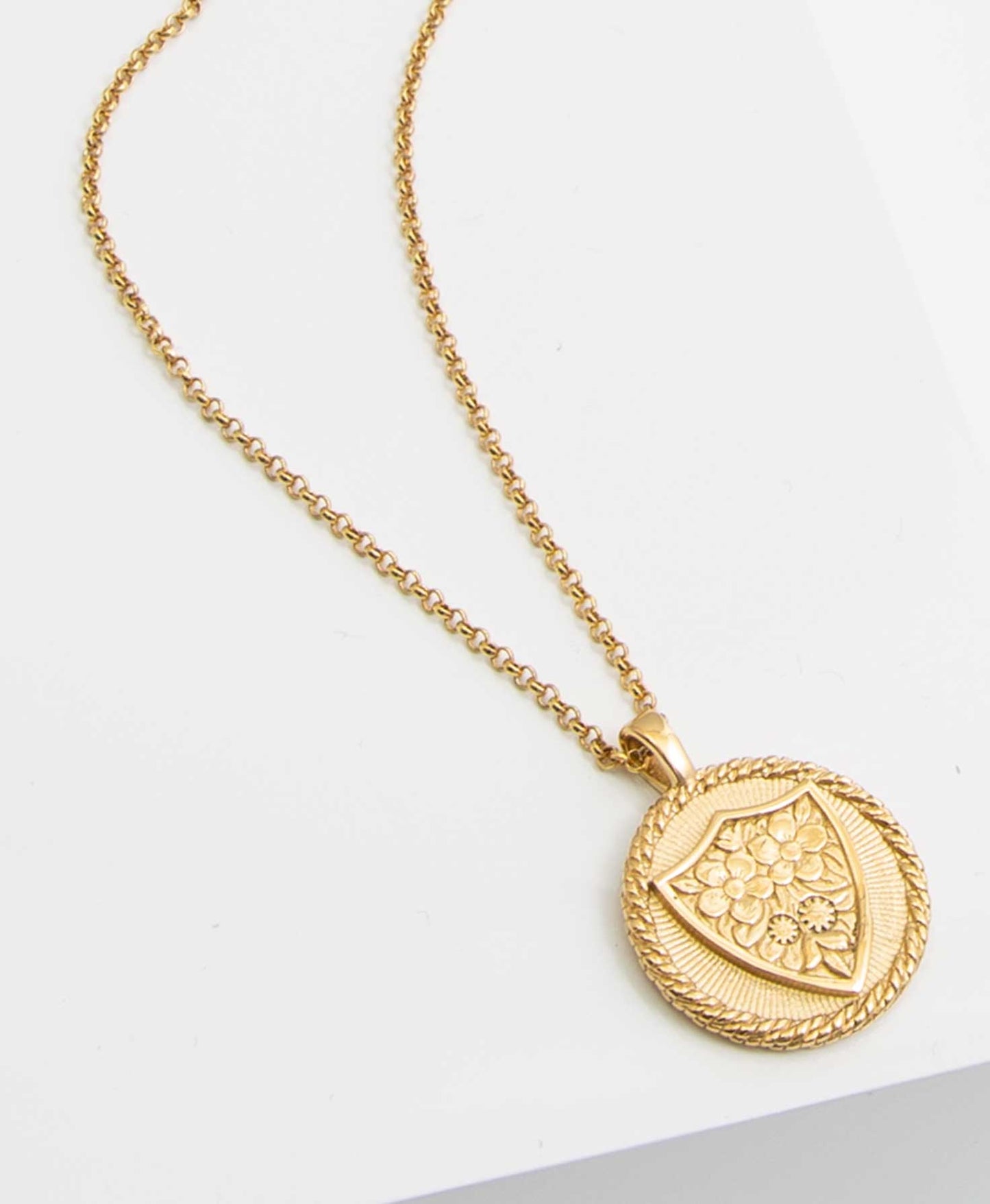 The Boundless Faith Necklace is a gold chain necklace with a circular gold pendant. The pendant has a gold vine border. In the center is a shield shape filled with golden flowers.