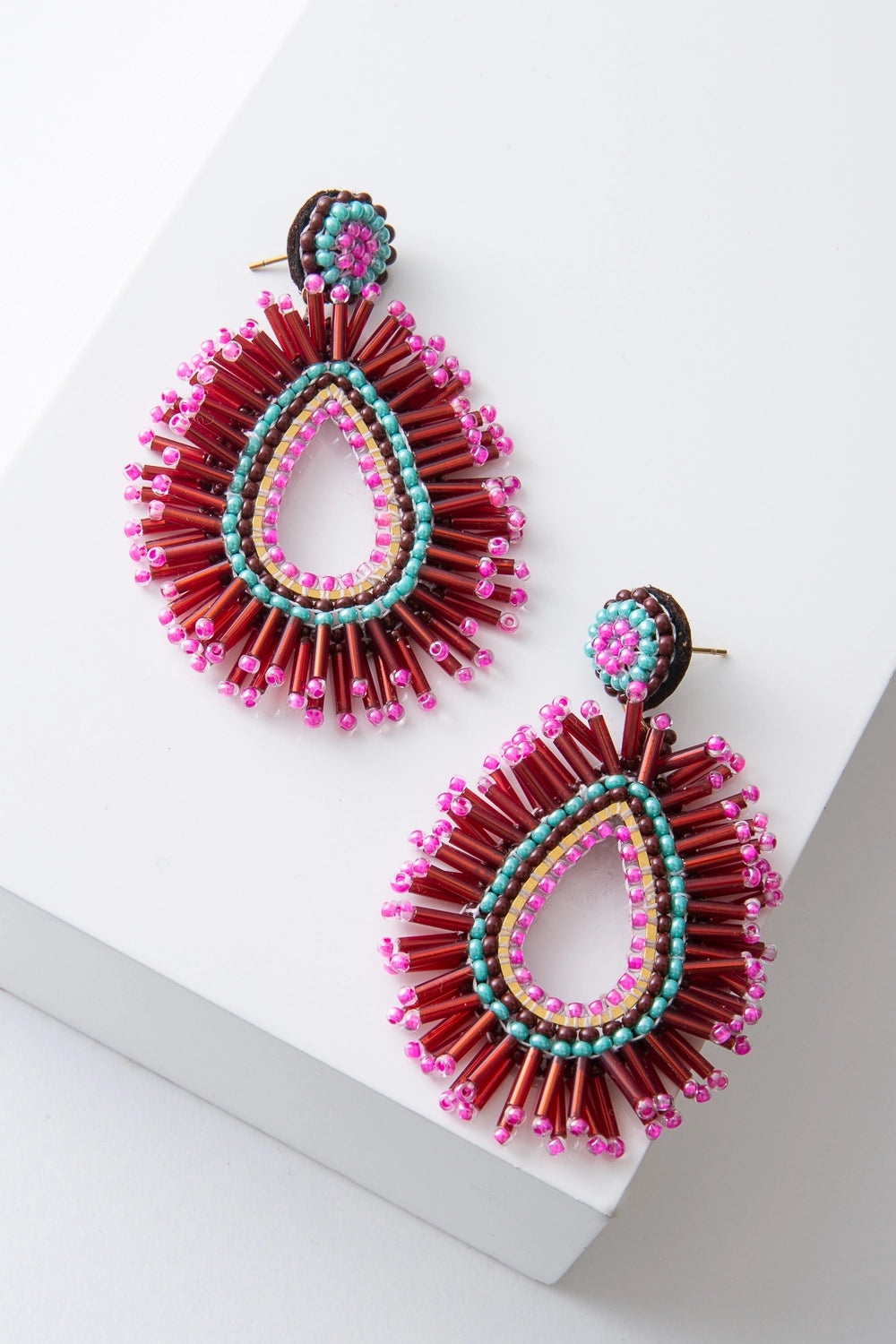These are brightly colored teardrop-style earrings that are open at the center. The ear post is adorned with aqua, brown, and hot pink beads. Hanging down from the post is a large teardrop composed of layers of hot pink, yellow, brown, and aqua beads. Around the exterior of the teardrop, there is a beaded fringe composed of tubular red beads accented with hot pink seed beads.