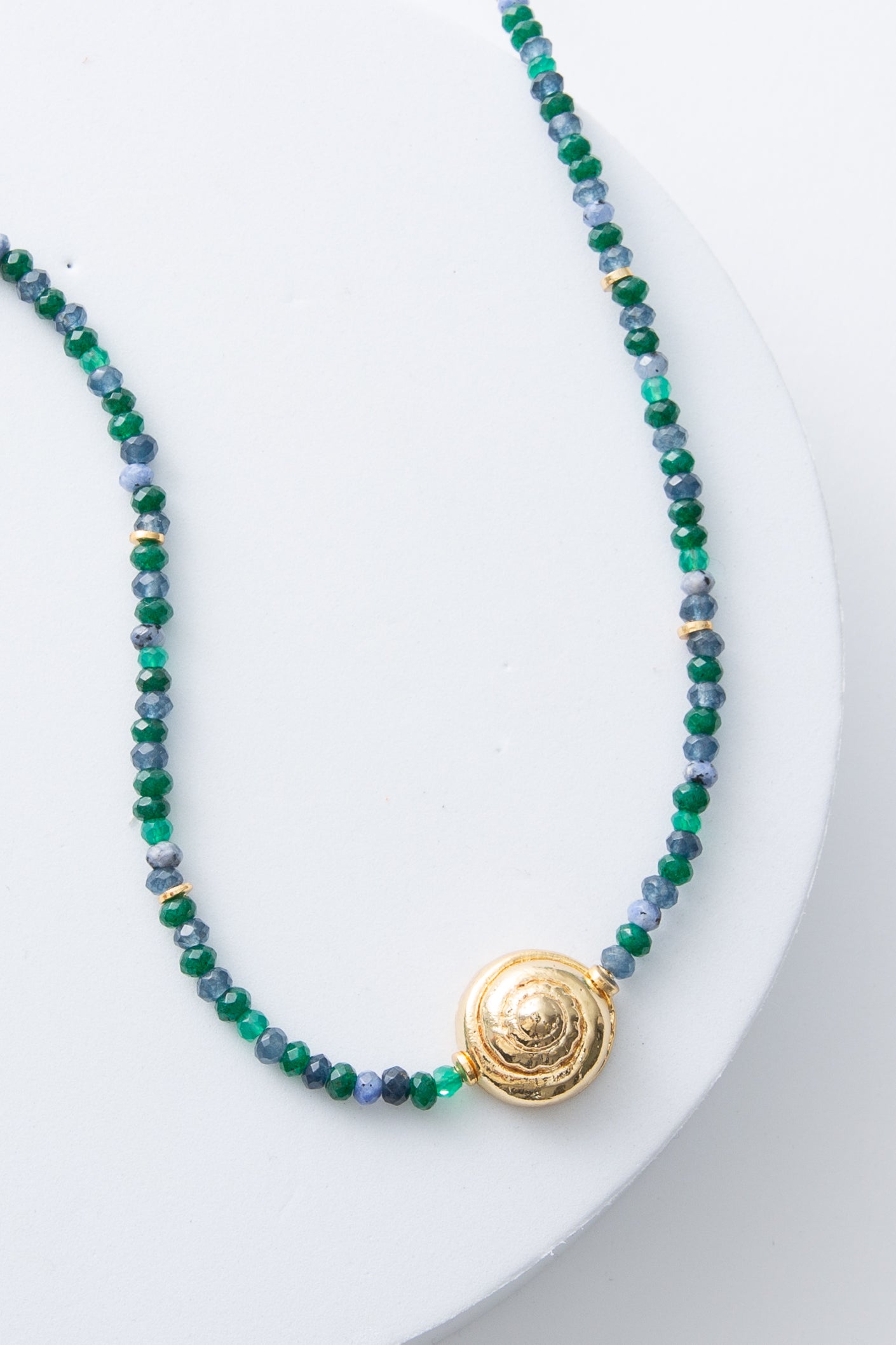 This is a single-strand necklace composed of small faceted beads made of agate stone. The stones are many shades of blue and green, creating an ocean-inspired look. In the center of the necklace there is a gold medal pendant in the shape of a spiraling snail shell.