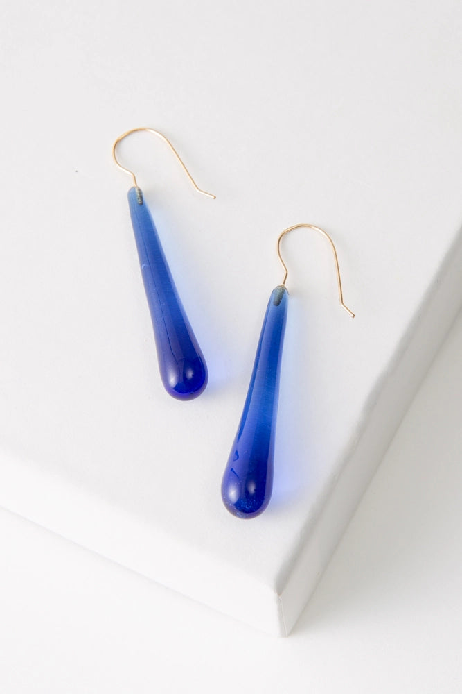 The Cenote Glass Earrings are long teardrop shaped earrings composed of translucent dark blue glass. The earrings have gold ear hooks.