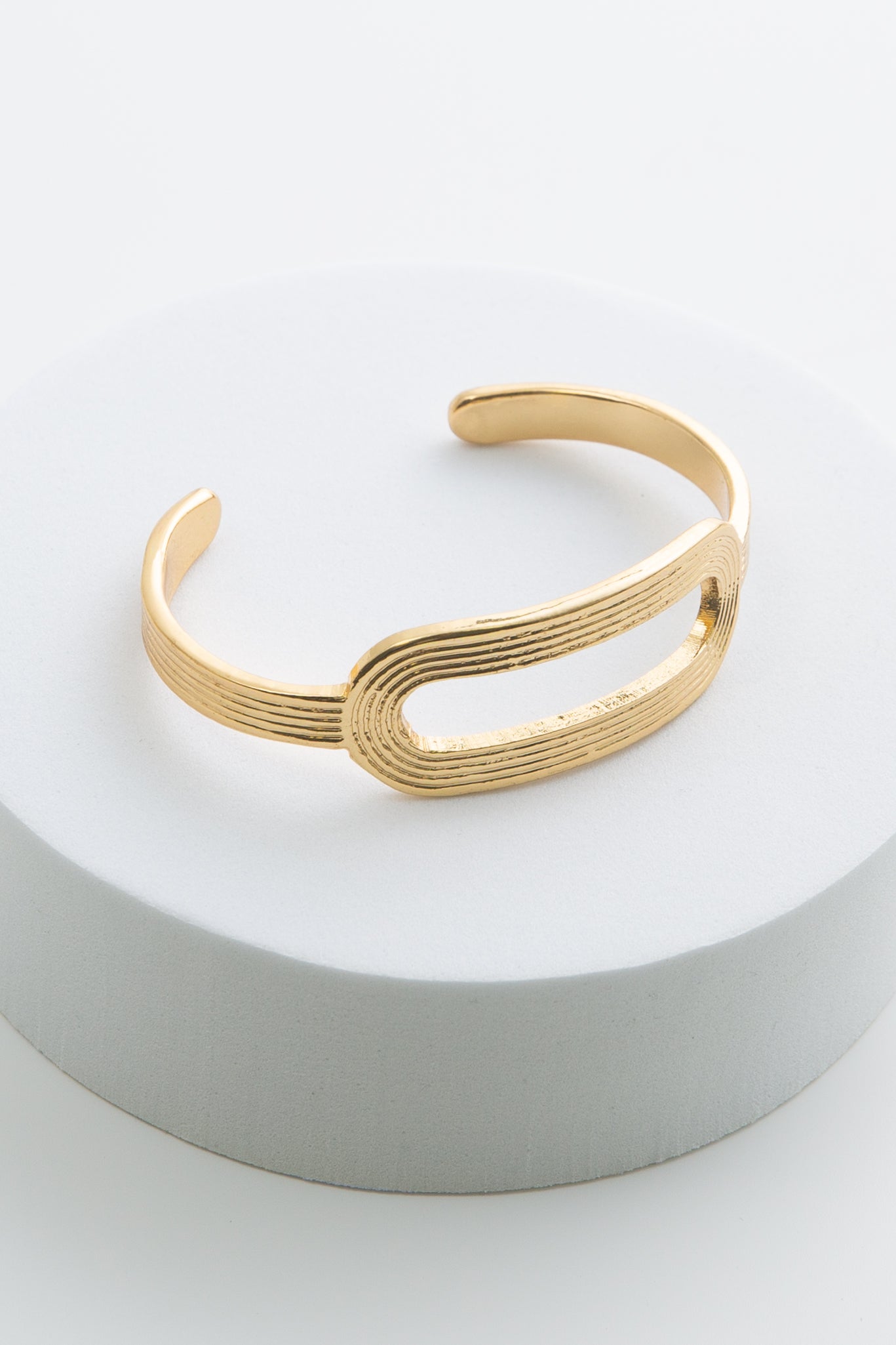The Circuit Cuff is a thin gold metal bracelet with an opening at the back where the bracelet can be slid onto the wrist. In the center of the bracelet is a horizontal oval made from the same gold metal that is hollow in the middle. There are horizontal lines etched all along the metal, giving the bracelet a ribbed look.