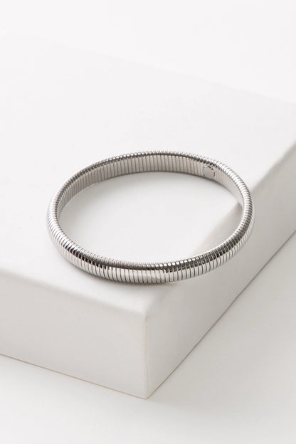 The Cobra Bracelet is a minimalist cuff composed entirely of shiny silver metal. The thin cuff is ribbed all the way around, giving it a coiled look.