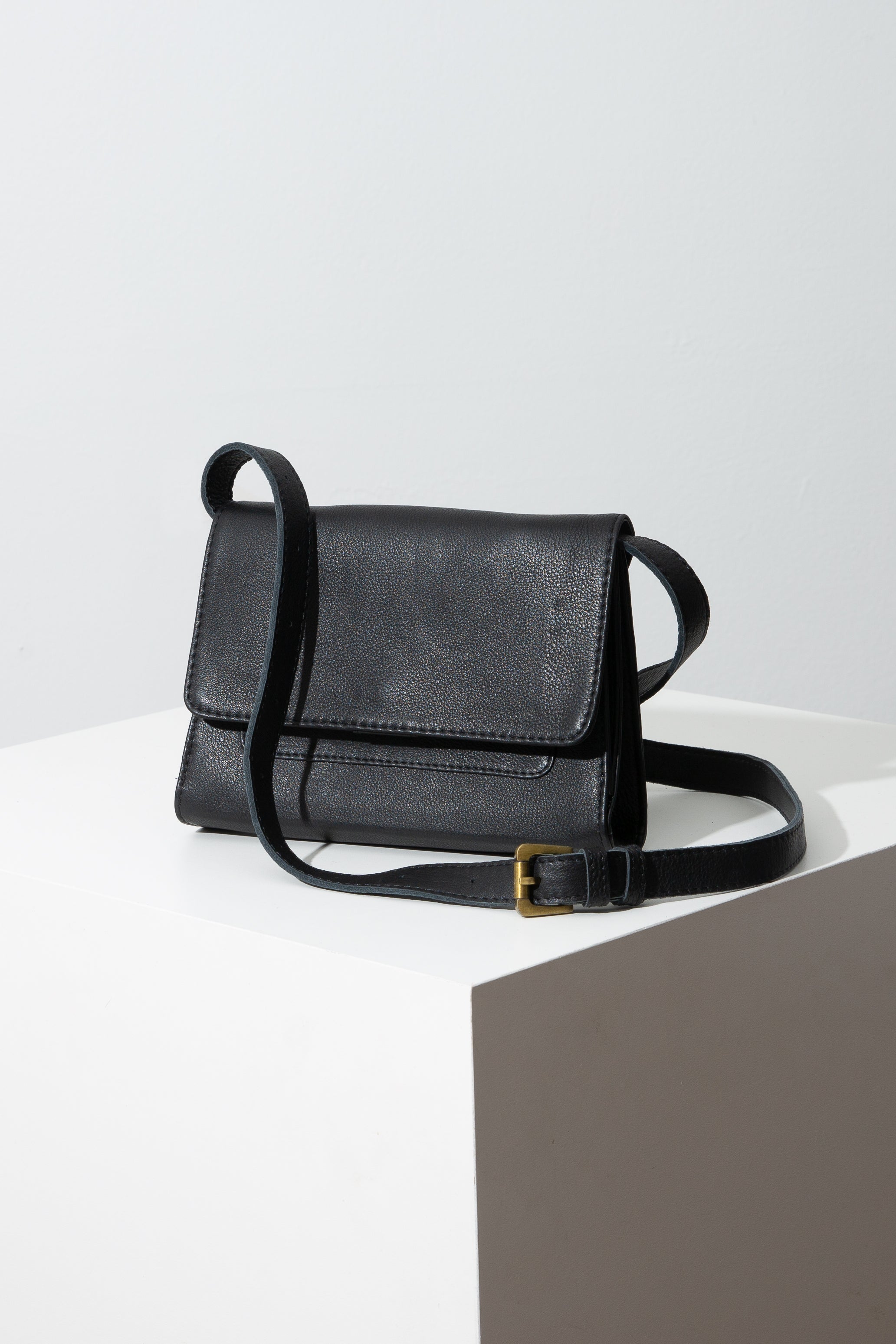 This is a rectangular bag that is slightly wider than it is tall. It is composed entirely of black leather. It has a flap that folds over the front of the bag and clasps shut. The bag can be worn several different ways. Here, an adjustable black leather strap is attached to the top corners of the bag.
