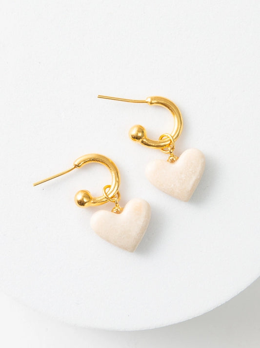 The Convertible Heart Huggies are small gold hoop earrings. There is a cream marble heart charm dangling from the hoops. Each hoop has a gold ball at the end to hold the charm in place. When the earrings are not being worn, the heart charms can slide off for a different look.