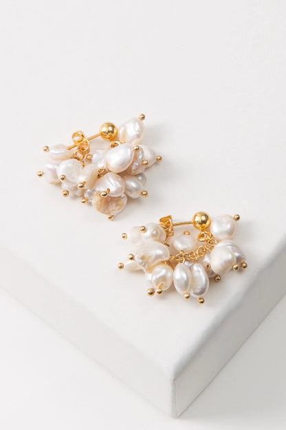 The Convertible Pearl Cluster Earrings are composed of round gold studs with a chain of dangling pearls attached to them. In this image, the earrings are shown with the pearls hooked to the back of the earrings, forming a small hoop. The pearls have a natural look and vary in shape and color.