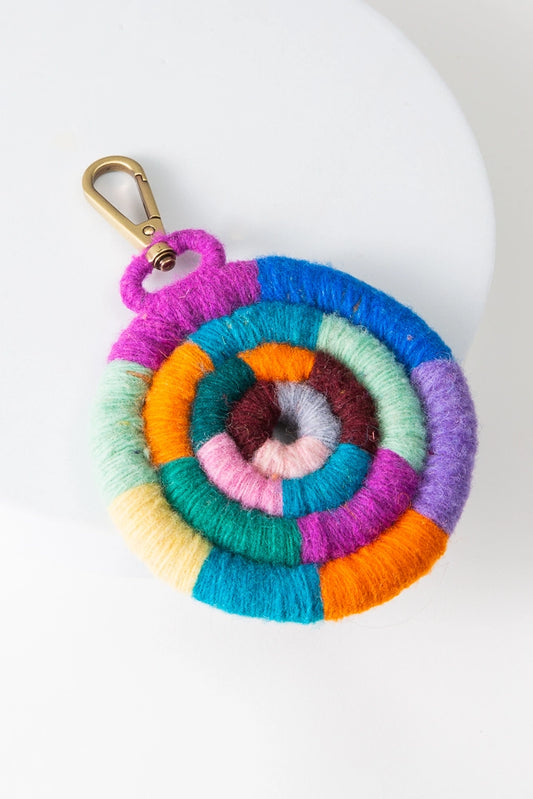 The Cookie Bag Charm is composed of multi-colored threads wrapped around a round cord. The cord is spiraled in on itself to form a round spiral shape. The threads are arranged in blocks of color and include magenta, blue, purple, orange, yellow, green, and crimson. The charm is attached to a gold clasp.