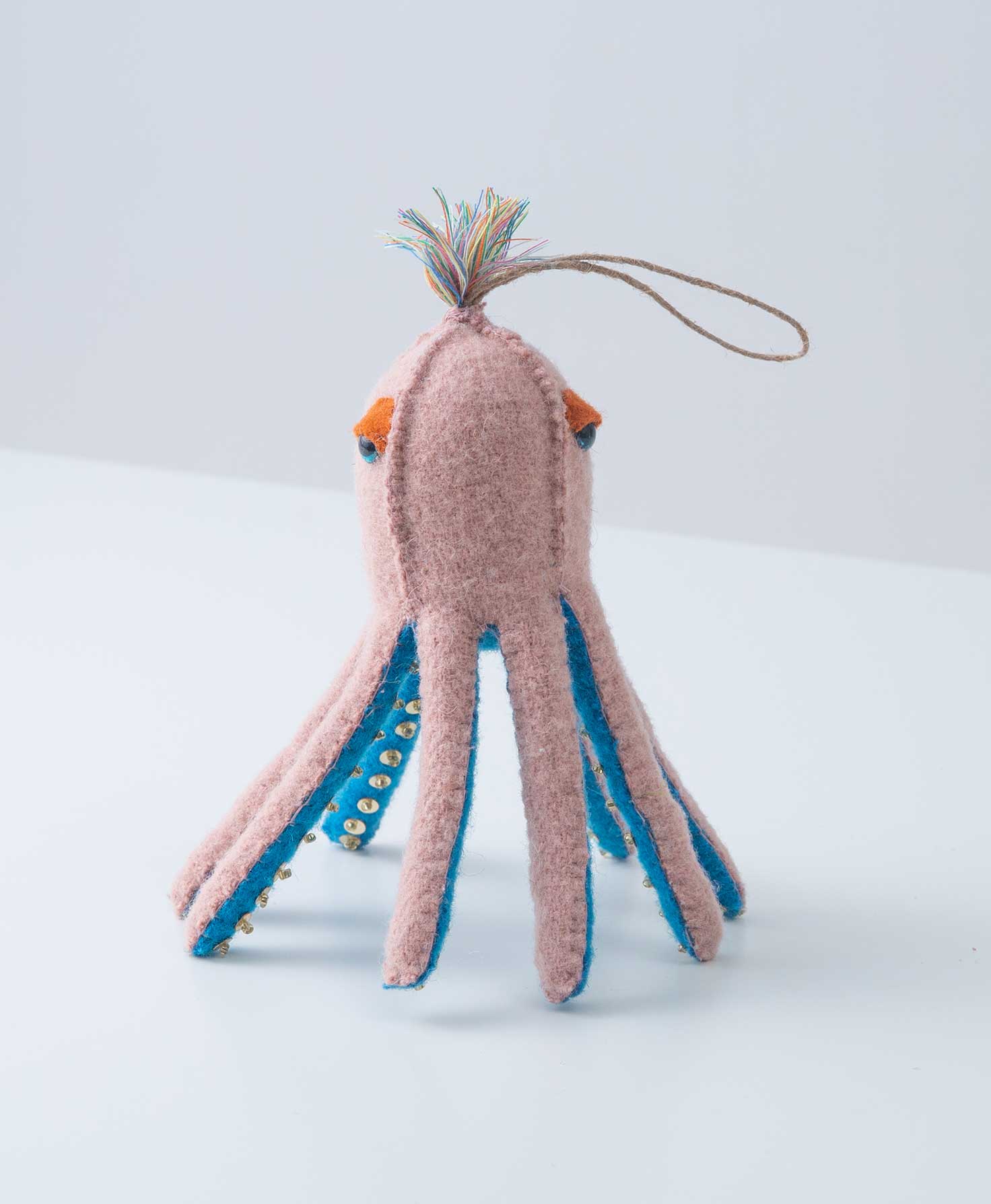 The Cosmic Octopus Ornament is composed of pink wool in the shape of a 3d octopus. The underside of each tentacle is blue wool with tiny beads stitched along it to give the look of tentacles. The octopus has shiny eyes and a tassel at the top of its head. There is a cord loop at the top, allowing the ornament to be hung.