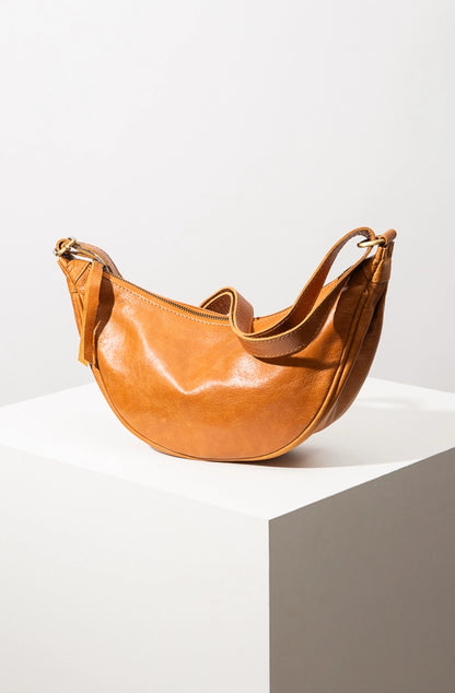 The Crescent Sling Bag is a half-moon shaped bag composed of glossy caramel-colored leather. The bag has a zipper closure with a leather tassel and an adjustable leather shoulder strap.