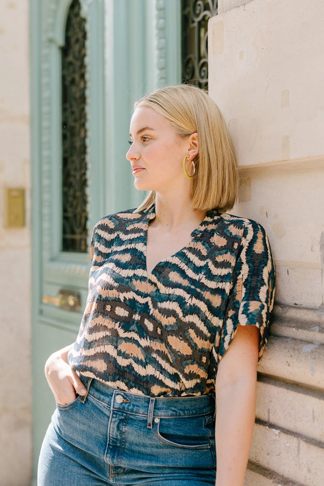 The Oversized Boxy Top in Vinita Sky is a short-sleeve blouse composed of a flowy rayon fabric. The fabric features a vibrant teal, cream, and tan graphic print that is reminiscent of a kaleidoscope pattern. The top has a v-neck and is pictured tucked into jeans.