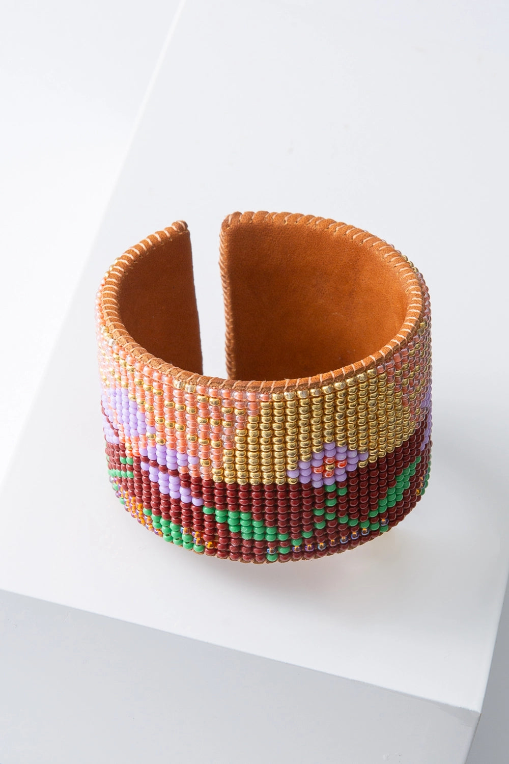 This is a wide, flexible beaded cuff. Soft leather is stitched with hundreds of tiny glass beads to form a desert landscape pattern. The background of the cuff is composed of rust, gold and pink beads that resemble a sunset. Lavender and green beads form flowers and leaves that form the foreground of the landscape.
