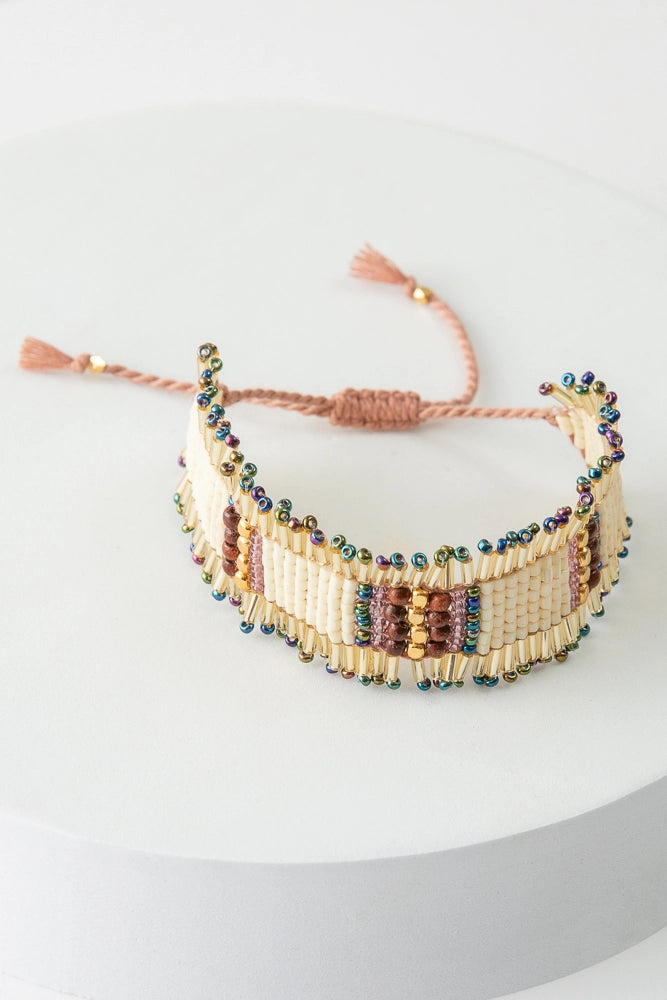 The Diwali Bracelet is a beaded bracelet composed of blocks of white beads that alternate with rows of green, pink, wood, and gold beads. The bracelet has a fringed gold beaded border. It has a braided string closure that can be tightened by pulling the ends apart.