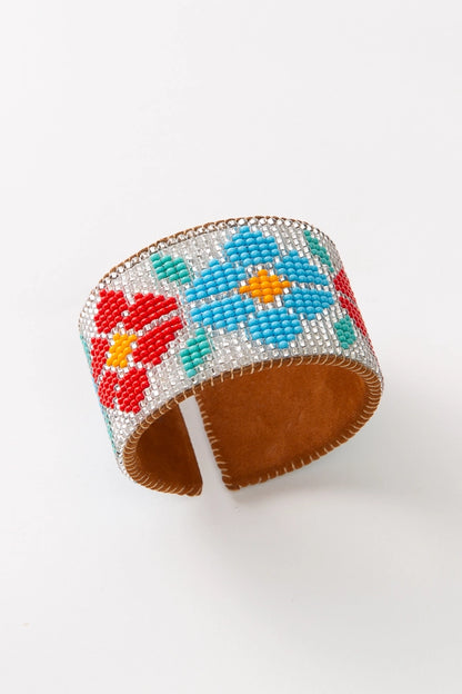 The Florecitas Cuff is a wide, flexible beaded cuff. Soft leather is stitched with hundreds of tiny glass beads to form a large floral pattern. The background of the cuff is composed of clear beads, and the flowers are red and light blue with mint green leaves.