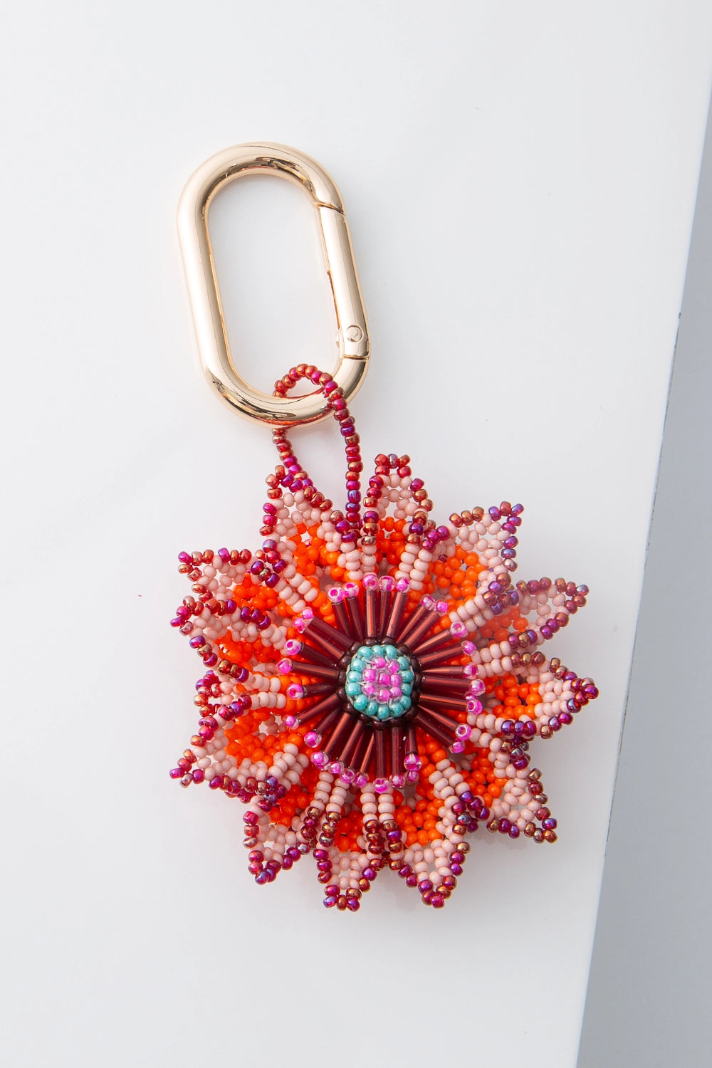 This is floral beaded charm that hangs from a brass oval keyring. The flower petals have a pointy shape and are composed entirely of tiny glass beads in shades of blush, magenta, orange, and red. The center of the flower is composed of tube-shaped red beads, with a circle of aqua and pink beads in the very center.