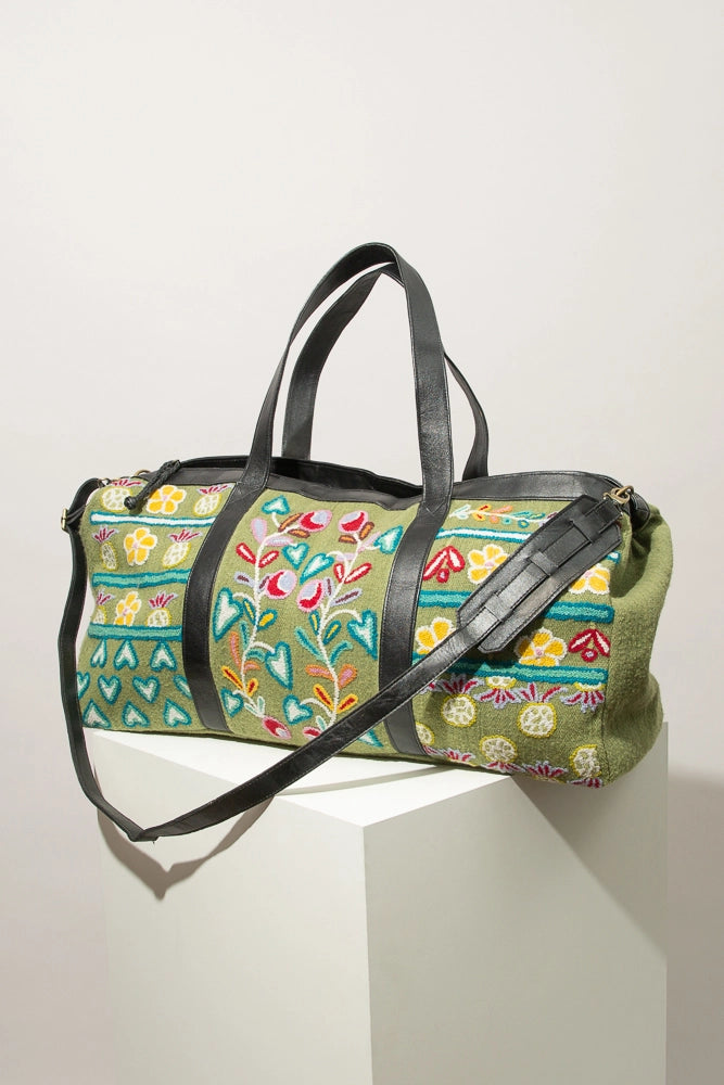 The Flourish Weekender is a large duffel-style bag composed of light green wool. Botanical motifs are embroidered all over the front of the bag with yellow, teal, red, and orange wool. The bag has two black leather handles as well as a long, removable black leather strap. There is a zipper closure at the top outlined in black leather.