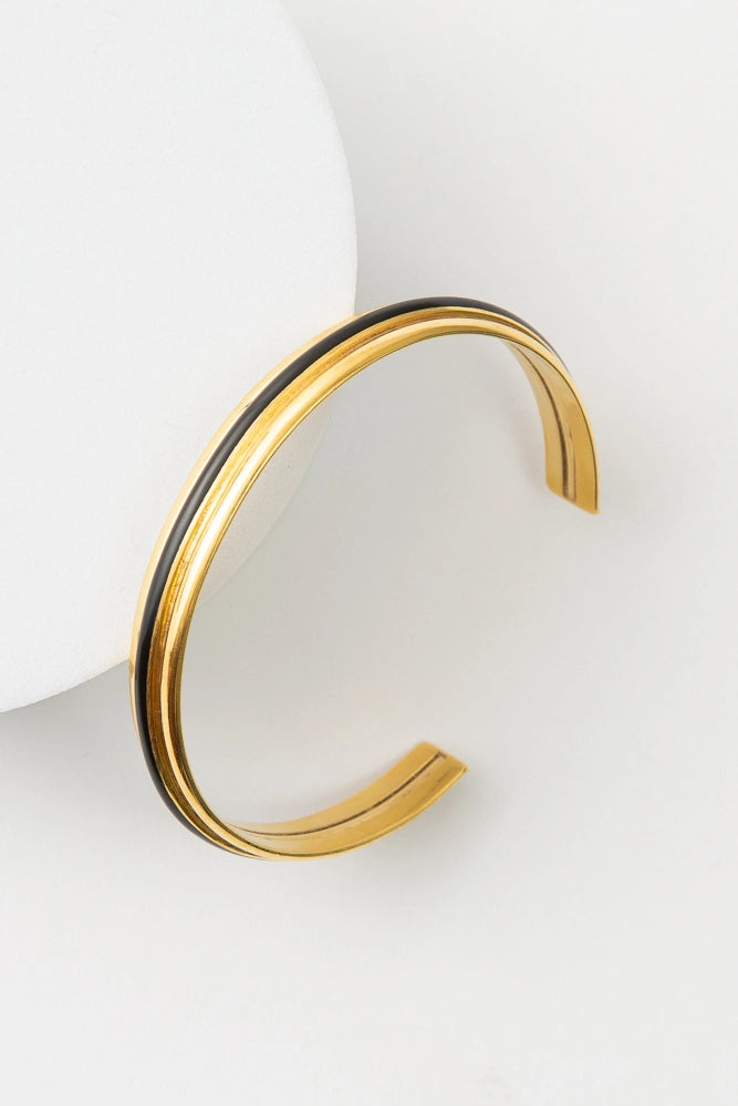 The Franco Cuff is a thin gold cuff that is open in the back, allowing it to be slid onto the arm. There is a narrow band of black resin stretching around the center of the cuff.