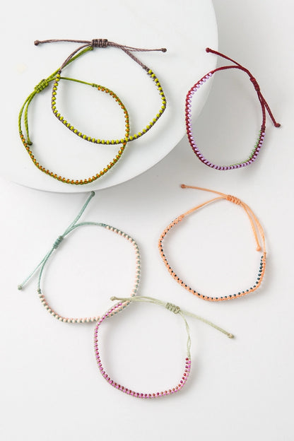 The six bracelet options available in the Friendship Bracelet Bar are pictured. Each bracelet is a different color, but they are all thin, single-strand thread bracelets embellished with dainty glass beads. They can be tightened by pulling on the ends of the thread.