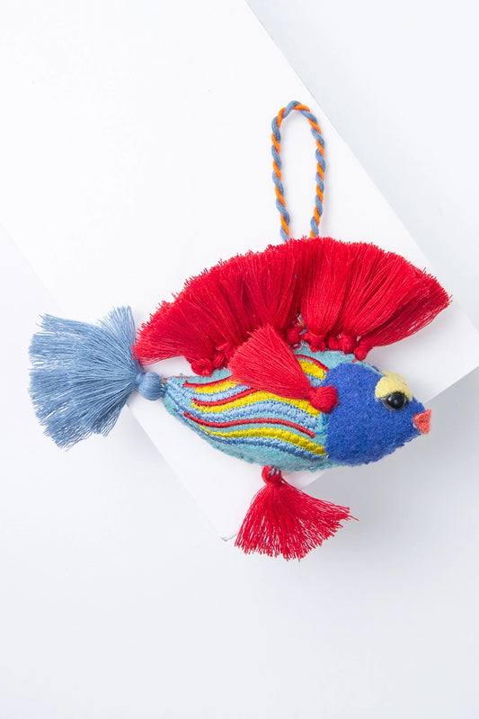 The Frisky Fish ornament is a bright wool ornament in the shape of a tropical fish. It has a light blue body with yellow, blue, and red stripes curving down it, and a dark blue head with black eyes and a peach mouth. The fish has bright red tassels all along its back. It also has small red tassels on its side and bottom to serve as fins, and a light blue tassel at its tail. The ornament has a woven orange and blue loop at the top, allowing it to be hung.