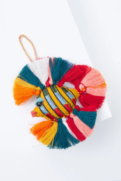 The Giddy Goldfish ornament is composed of gold wool sewn into the shape of a fish. There are stripes of green, blue, and red running vertically along the body. The face is finished with a pink mouth and shiny black eye. Brightly colored tassels in shades of orange, yellow, teal, white, red, and pink are attached along the top, back and bottom of the fish to form fins. There is a woven loop at the top of the ornament where it can be hung.