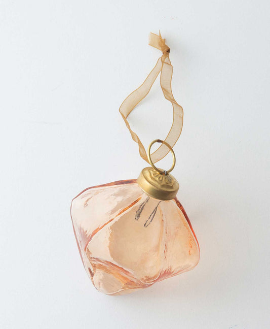 The Glass Bulb Ornament in peach is composed of transparent peach colored glass in the shape of a three dimensional diamond. the interior is hollow. There is a piece of brass at the top that has a peach ribbon loop attached to it, allowing the ornament to be hung.