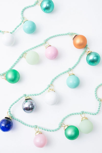 The Glass Globe Garland is composed of a twisted mint green cord with glass ball ornaments tied to it every few inches. Some of the ornaments have a matte finish, while others have a metallic finish. They are shades of aqua orange, green, mint, white, silver, pink, and blue. The hardware at the top of the ornaments is gold metal.