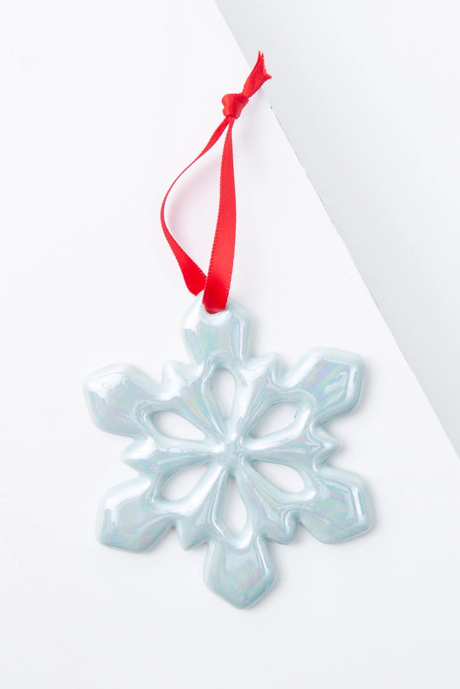 The Glazed Snowflake Ornament is a snowflake shaped ornament made of white porcelain with a shiny, iridescent finish. There is a red ribbon tied to the top of the ornament, allowing it to be hung.