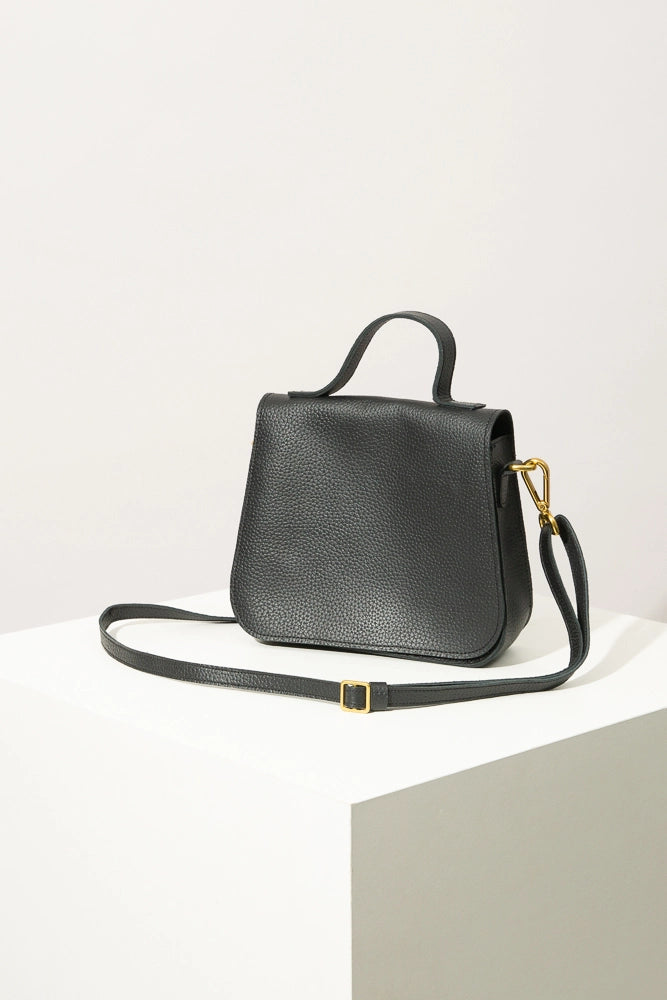 The Go-Getter Bag is a small, roughly square handbag with rounded corners. It is composed of black leather and has a flap that folds over the front of the bag. There is a short leather handle on top of the bag, and an adjustable and removable long strap attached to the sides. The strap is attached with gold hardware.