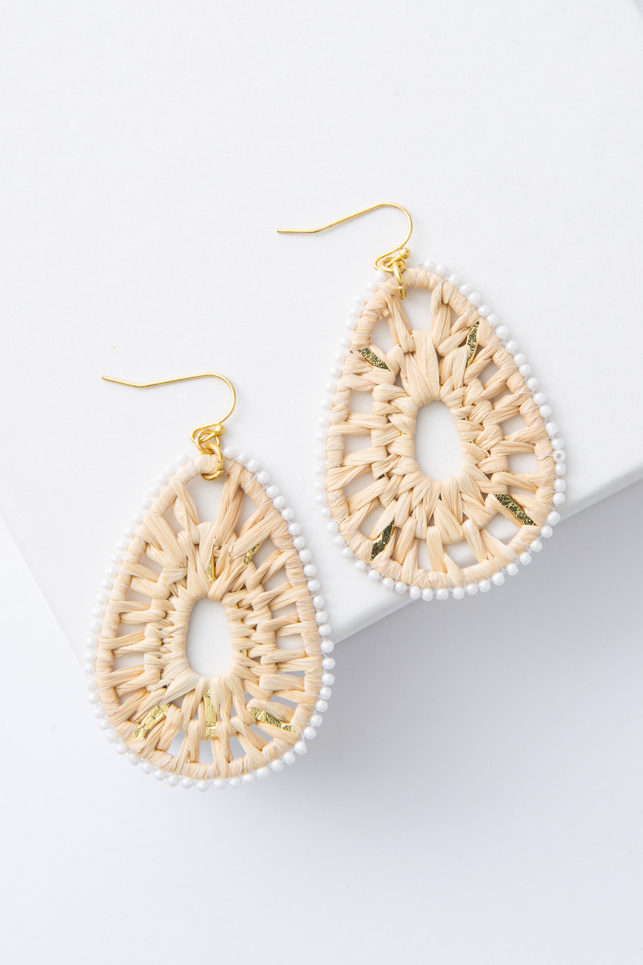 The Goa Earrings are tear-drop shaped earrings composed of woven raffia with an outline of white glass beads. The raffia has a lace-like pattern and is open in the center. The large teardrops hang from brass ear wires.