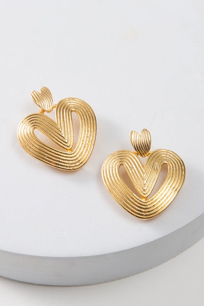 The Golden Palmier Earrings are post-style earrings composed of thin rods of brass that have been stacked and molded into a heart shape. The heart is open in the center. The ear posts themselves are smaller brass hearts that are fused to the larger hearts below.