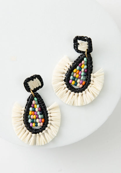 The Gulal Earrings are large teardrop chandelier earrings. The diamond shaped posts are wrapped in black and white raffia. A teardrop shape hangs down below the posts. The exterior rim is wrapped in black raffia, and there is white raffia fringe along the bottom. The interior is composed of rainbow colored glass beads.