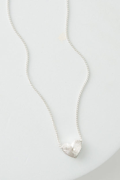 The Heart Swell Necklace has a dainty silver chain. The chain is attached to either side of a small silver heart pendant. The heart has many lines etched into it emanating from the center, giving it a starburst appearance.