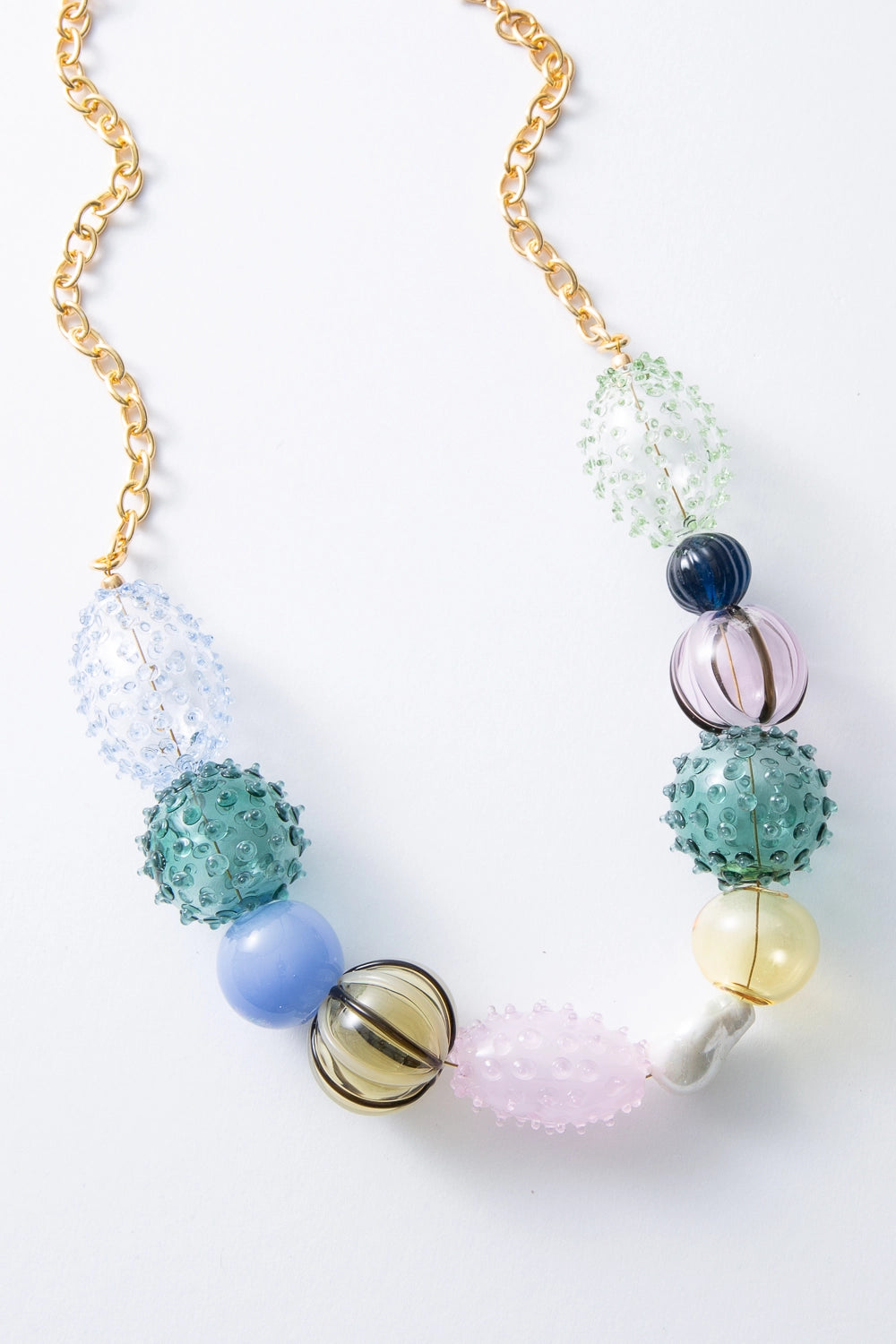 The Iris Glass Blown Necklace has a gold link chain at the top of the necklace, and the bottom is strung with three-dimensional glass beads in various shapes, textures, and colors. There are oval-shaped and spherical beads covered in a spiky texture, smooth spherical beads, and ribbed spherical beads. The colors of the beads include teal, navy, pink, yellow, and mint. Among the glass beads, there is a natural pearl with an irregular, organic shape.