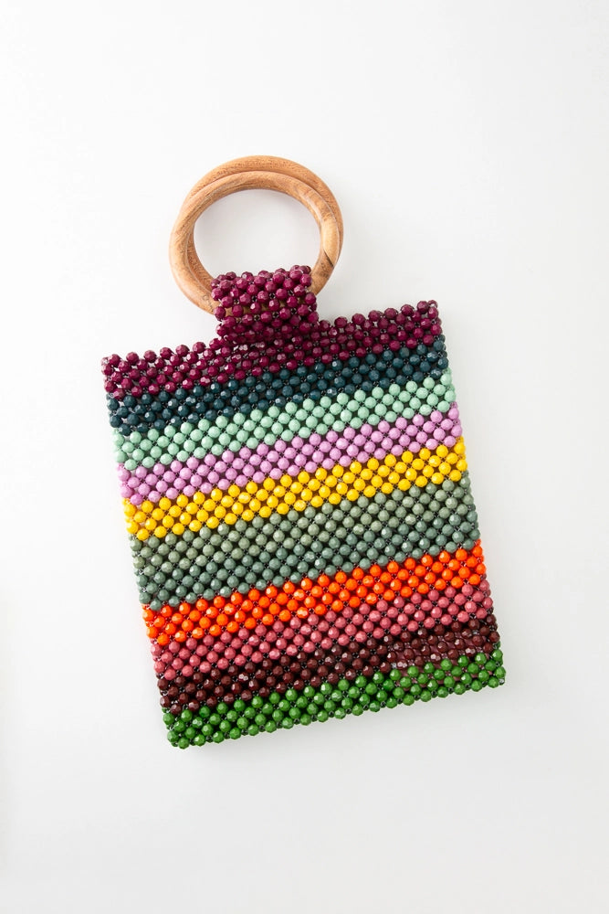 The Je T'aime Bag is a rectangular bag that is longer than it is wide. It is composed entirely of stripes of brightly colored beads in shades of green, burgundy, teal, mint, pink, yellow, and orange. There are two round wooden handles attached to the top of the bag.