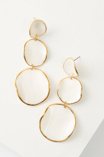 The Le Fantasia Earrings are chandelier drop earrings composed of a stack of three white porcelain discs outlined in shining gold. The disc attached to the post is the smallest, and each disc below is a little larger. The discs are not flat and have curves that give them a more organic look.