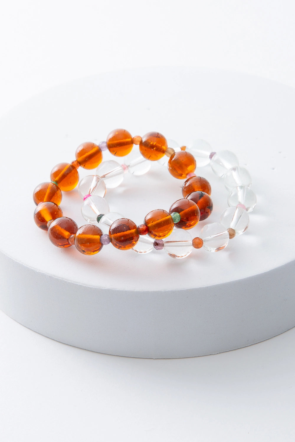 The two bracelets included in the Lucid Glass Bracelet Set are pictured together. Both are single-strand bracelets composed of spherical glass beads, with smaller semi-precious stone beads in a variety of colors in between. One bracelet is composed of clear glass spherical beads. The other bracelet is composed of translucent amber colored glass beads.