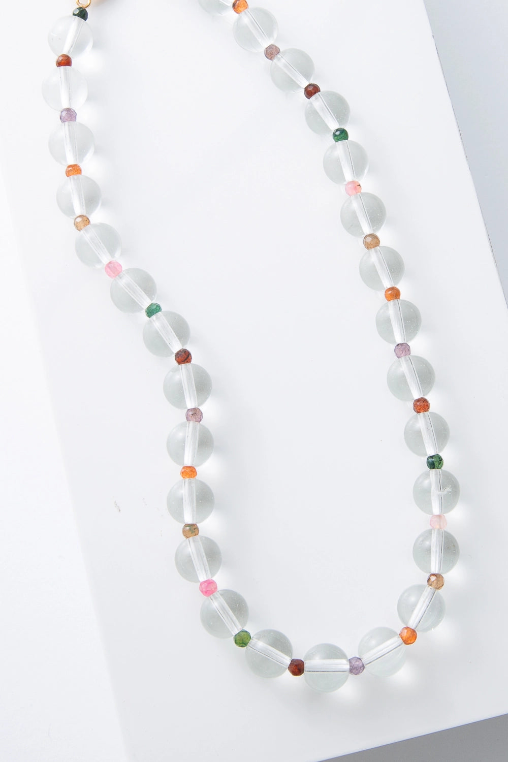 This is a single-strand necklace composed of large, clear glass spherical beads strung all along the length of the necklace. In between each pair of clear beads there is a small faceted bead made of a different colored semi-precious stone, creating a rainbow look. 