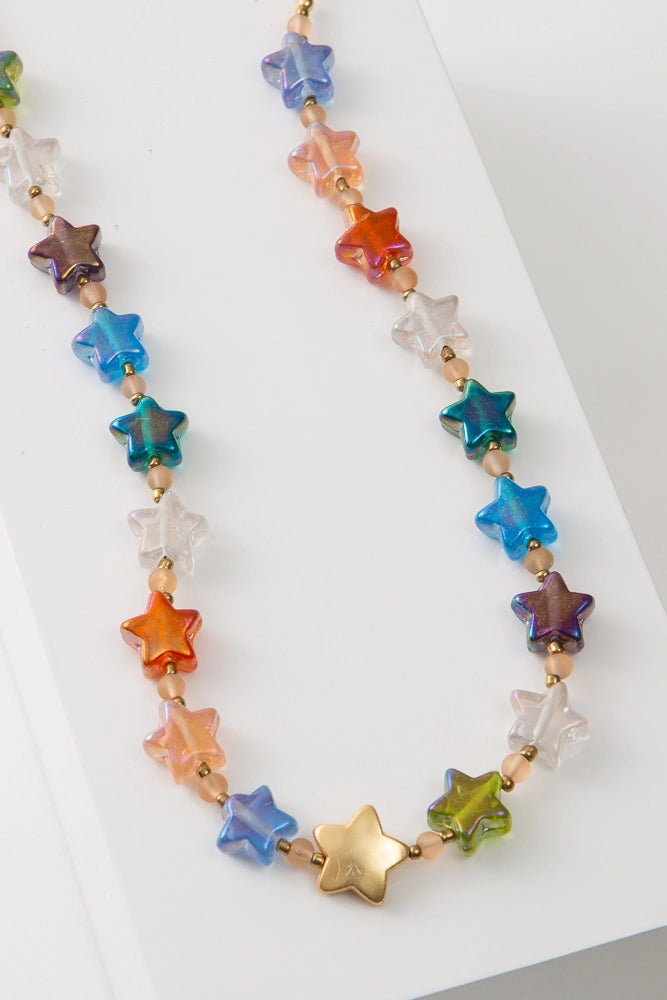 The Lucky Star Glass Necklace is composed of a single strand of glass star beads. The stars are shades of purple, white, green, red, and blue. There is a larger brass star at the front of the necklace. Small pink spherical glass beads and brass seed beads are strung between the stars.