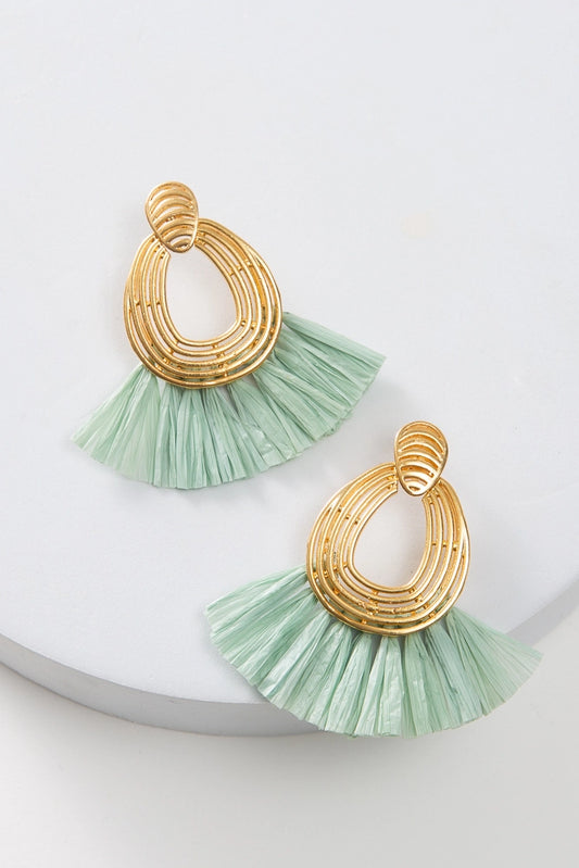 The Lux Raffia Earrings are post-style hoop earrings. The hoop is composed of thin rods of brass molded into an oval shape that is open in the center. Mint colored raffia fringe fans out from the bottom of the hoop.