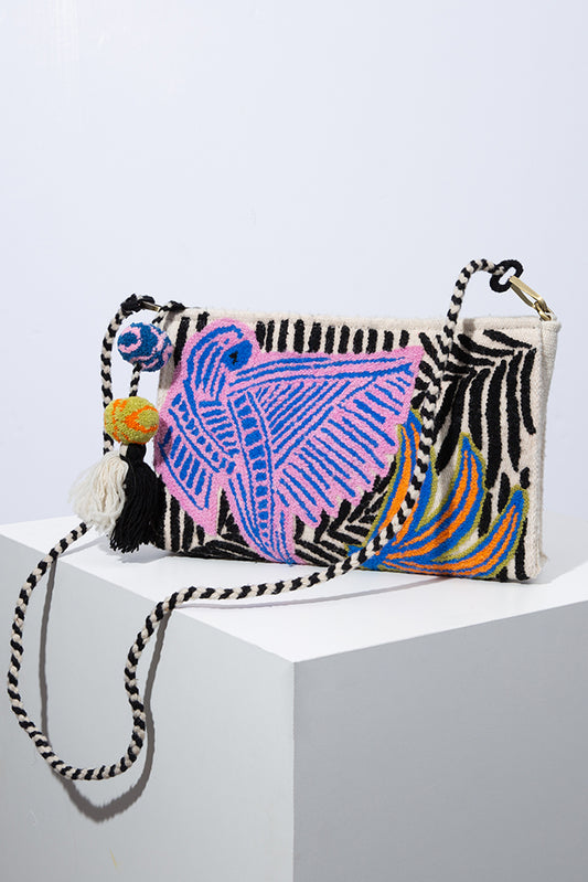 The Macaw Crossbody is a rectangular bag that is wider than it is tall. It is composed of white wool with palm frond designs embroidered in black wool across the bag. A large abstract macaw design is stitched in blue and pink on the front of the bag. The bag has a zipper closure. There is a long strap composed of black and white wool attached to either corner of the bag.