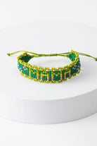 The Mama Beaded Bracelet is an adjustable thread bracelet featuring a band of green glass beads. Within the band, the word "Mama" is formed with lime, yellow, and pink glass beads. A row of lime glass beads forms a border around the bracelet. A lime thread closure at the back of the bracelet can be adjusted by pulling on both ends of the thread.